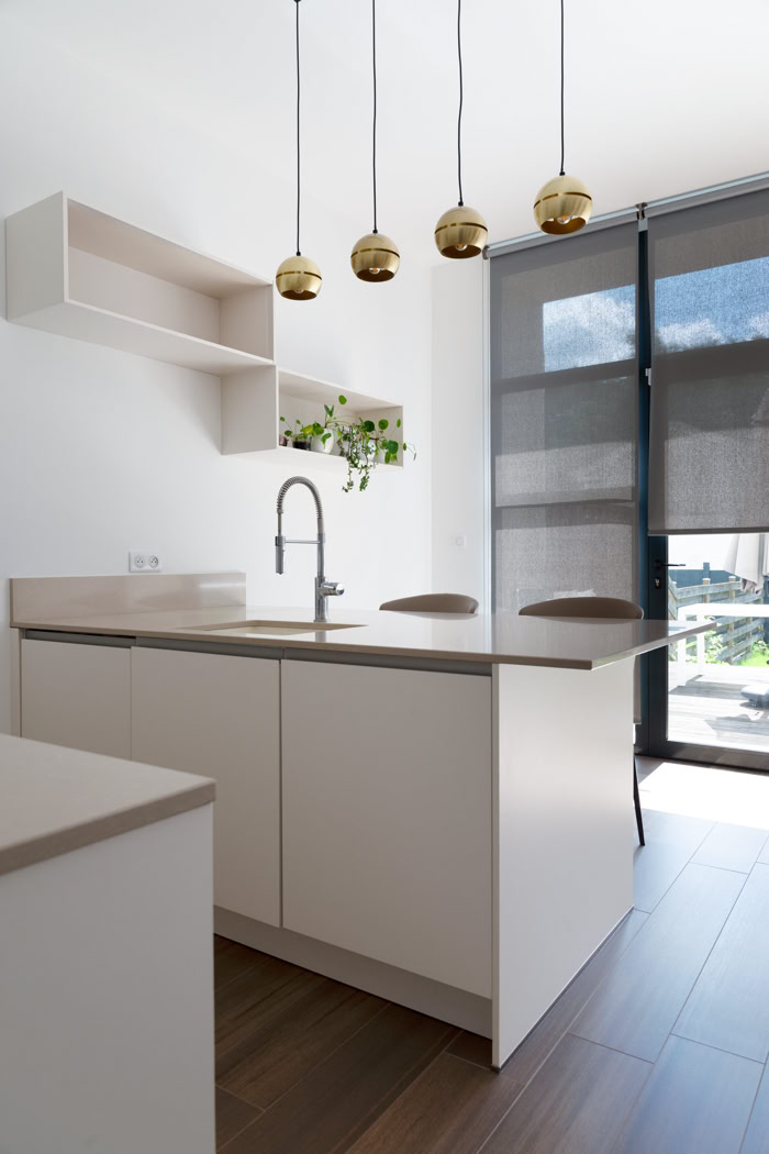 Cuisine blanche minimaliste avec îlot central, plan de travail beige et suspensions dorées, réalisation d'architecte d'intérieur à Thionville, Moselle Est.