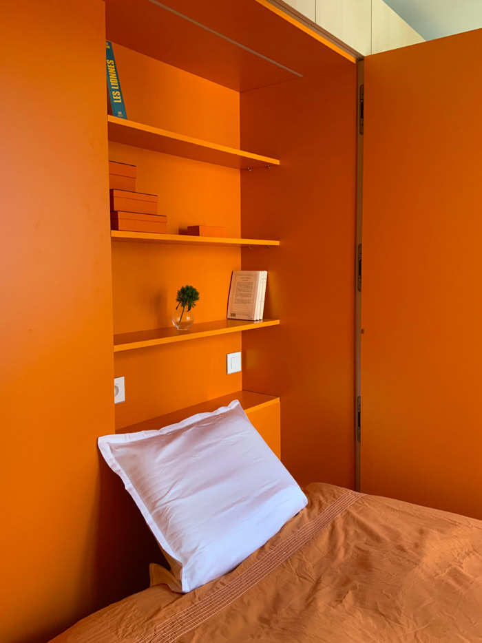 Zoom sur l'espace nuit orange vif avec tête de lit et étagères intégrées dans un studio de 17 m².